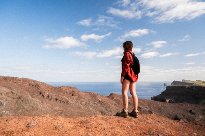 A girl with a red shirt and black backpack is standing on rocks and looking at the sea