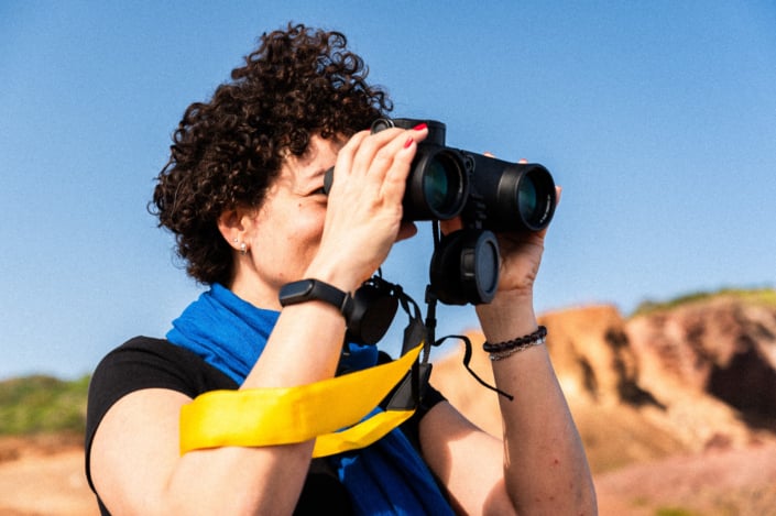 A woman with short curly hairs and a blue scarf is looking through binoculars