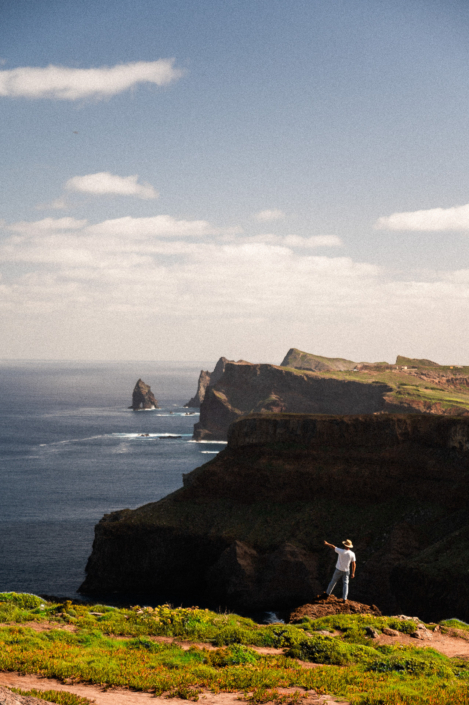 A male digital nomad with a white shirt and a hat pointing in the distance, standing in front of the rocky coastal landscape of Madeira island