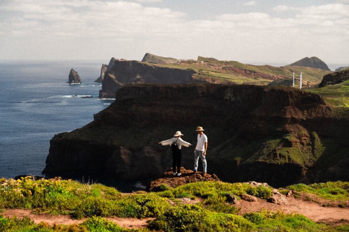 A digital nomad couple standing on a cliff, she is spreading her arms, he is standing in front of her, magnificent landscape in the background